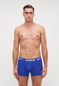 Boxeri Nike albaștri cu o talie albă cu marcă, având o croială strânsă și un material neted, elastic. Fără modele sau detalii suplimentare.