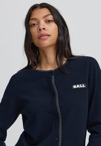 Cárdigan de punto en color marino con cuello redondo, tapeta de botones grises y una etiqueta que dice "BALL" en el lado izquierdo del pecho. Textura suave y forma ajustada.