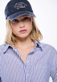 Cappello della marina con testo ricamato "SLOW SUNDAYS COFFEE CLUB," indossato con una camicia a righe blu e bianche con taschino sul petto.