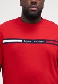 Raudona medvilninė T-maišelis su horizontalia mėlyna ir balta juostele bei "TOMMY HILFIGER" tekstu, trumpomis rankovėmis ir apvaliu kaklu.