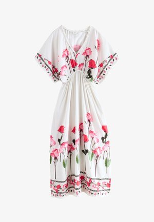 Mujer descalza de pie en escalones exteriores vistiendo un vestido largo blanco con estampados florales rosas y mangas sueltas, junto a vegetación y paneles de madera.