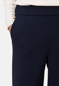 Personne portant un pantalon bleu marine foncé avec une poche latérale, une main partiellement à l'intérieur de la poche, et un haut à manches longues beige clair.