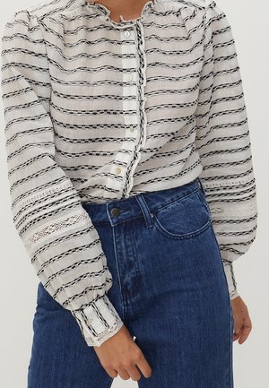 White blouse with black horizontal stripes, round neckline, button front, and puffed sleeves featuring intricate lace accents. Paired with blue jeans.