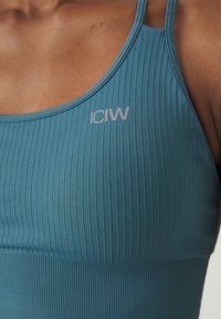 Sostenedor deportivo de canalé en color verde azulado con tirantes ajustables. Presenta un pequeño logotipo gris "ICIW" en la parte superior izquierda. Tejido suave y elástico.