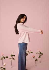 Frau im rosa gestreiften Hemd und Jeans streckt die Arme seitlich aus, umgeben von rosa Rosen in Glasvasen vor rosa Hintergrund.