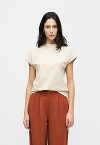Μπεζ βαμβακερό μπλουζάκι με κοντά, γυρισμένα μανίκια και στρογγυλή λαιμόκοψη, σε συνδυασμό με παντελόνια ψηλόμεσα σε σκουριαστού χρώματος με πιέτες.