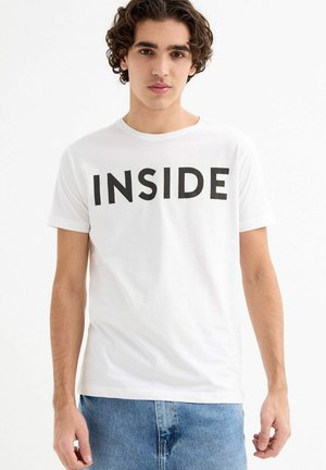 Joven con cabello rizado, vistiendo una camiseta blanca con el texto "INSIDE" y jeans azules, posando frente a un fondo blanco liso.