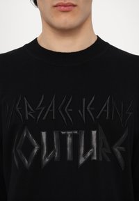 Juodas megztinis su iškeltu, tekstūriniu juodu logotipu "VERSACE JEANS COUTURE" drąsiu, kampuotu šriftu. Paprastas, apvalios kaklo iškirpimo dizainas.