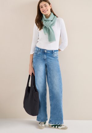 Femme souriante portant un haut blanc, un jean large bleu clair, une écharpe verte, des baskets, et tenant un sac fourre-tout noir sur un fond neutre.