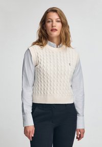 Elfenbeinfarbener, kabelgestrickter Strickwesten mit Rundhalsausschnitt und geripptem Saum, getragen über einem gestreiften blauen und weißen Hemd mit Kragen, kombiniert mit dunklen Hosen.