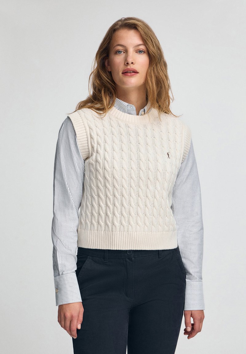 Elfenbeinfarbener, kabelgestrickter Strickwesten mit Rundhalsausschnitt und geripptem Saum, getragen über einem gestreiften blauen und weißen Hemd mit Kragen, kombiniert mit dunklen Hosen.