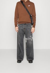 Hnědá mikina s přední kapsou a bílým logem, doplněná o oversized černé džíny s popraskanými detaily a třepenými okraji. Černé boty.