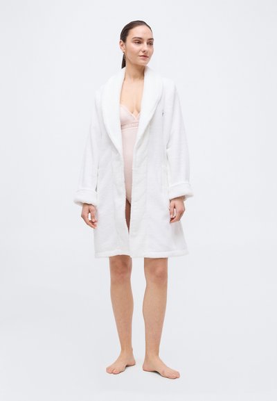 Γυναίκα όρθια, ξυπόλητη, φορώντας λευκή ρόμπα από απαλό ύφασμα πάνω από ένα ροζ δαντελωτό κορμάκι, με φόντο απλό και φωτεινό.
