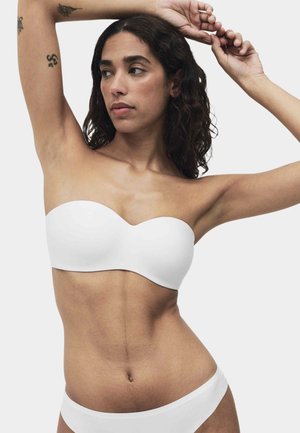 Kvinde med krøllet mørkt hår iført en hvid stropløs bh og matchende undertøj, løfter armene over hovedet og viser tatoveringer på armene.