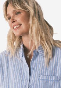 Jasnoniebieska koszula w paski z guzikami i kieszenią, miękka w dotyku, z klasycznym kołnierzykiem. Guziki są okrągłe, pasujące do materiału.