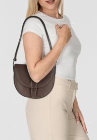 Braune Leder-Umhängetasche mit strukturiertem Finish, geschwungener Form und goldfarbener Schnallenakzent; getragen auf einer Schulter mit einer hellen Bluse.