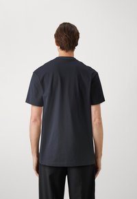 Tummansinisestä puuvillapaidasta, jossa on lyhyet hihat ja pyöreä kaula-aukko, on esitetty takaa. Paidassa on sileä pinta ja minimalistiinen muotoilu.