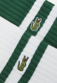 Toalla de algodón con franjas verdes y blancas, adornada con dos logotipos de cocodrilo bordados. Superficie texturizada con elementos acanalados.