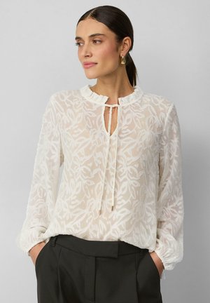 Witte, doorschijnende blouse met bloemstickers, gerimpelde halslijn en striksluiting. Lange mouwen met elastische manchetten; gecombineerd met zwarte broek.