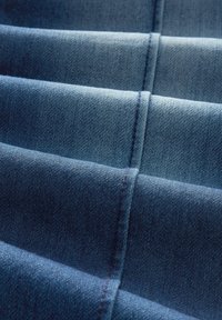 Gros plan sur un tissu en denim bleu plié montrant des coutures détaillées le long des plis, éclairé par une lumière douce et uniforme.