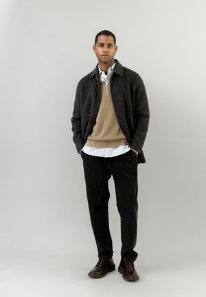 Mørk ulljakke over en beige genser, lagdelt med en hvitstripet skjorte, kombinert med mørke teksturerte bukser og brune skinnsko.