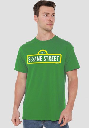 Green cotton t-shirt featuring "123 SESAME STREET" graphic in yellow and white, crew neck, and short sleeves. Smooth texture and relaxed fit.