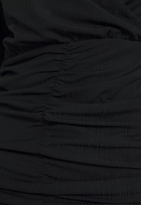 Tissu noir texturé avec des détails froncés, créant des plis verticaux. Le matériau est extensible et léger, améliorant la silhouette du vêtement.