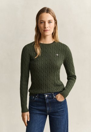 Vrouw met lang blond haar die een donkergroene kabelgebreide trui en blauwe jeans draagt, met één hand in de zak staand tegen een beige achtergrond.