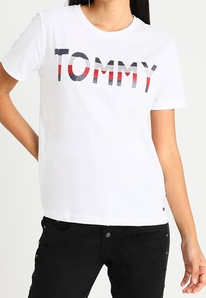 Frau trägt weißes T-Shirt mit kurzen Ärmeln und dem Schriftzug "TOMMY" in Schwarz, Weiß und Rot, kombiniert mit schwarzen Hosen, steht mit einer Hand in der Hüfte.