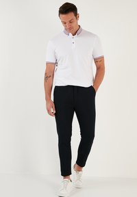 Valkoinen polo-paita, jossa on violetti kaulus ja hihansuut, yhdistettynä mustiin slim fit -henkisiin housuihin ja valkoisiin lenkkareihin. Malli seisoo, katsoo alas.