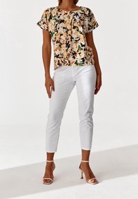Blusa a maniche corte con motivo floreale nei toni crema, verde e arancione, abbinata a pantaloni bianchi slim-fit e sandali con tacco nudo.