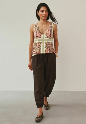 Frau, die ein ärmelloses, rosa gemustertes Oberteil, lockere dunkelbraune Hosen und schwarze, gewebte Ballerinas trägt, vor einem schlichten Hintergrund geht.