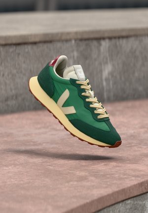 Zöld és krémszínű sneaker bézs cipőfűzőkkel, amint lebeg egy betonfelület fölött, oldal- és felülnézetben.