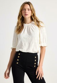 Lyshåret kvinde iført en hvid tekstureret bluse med pufærmer og højtsiddende sorte bukser med to vertikale rækker af guldknapper.