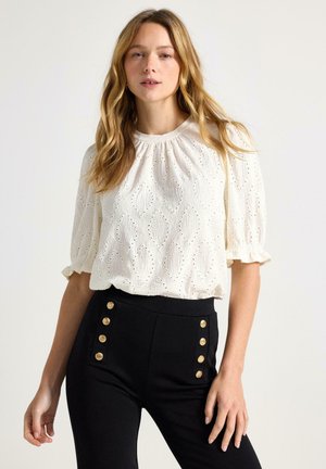 Lyshåret kvinde iført en hvid tekstureret bluse med pufærmer og højtsiddende sorte bukser med to vertikale rækker af guldknapper.