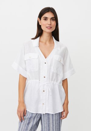 Kvinde iført en hvid kortærmet skjorte med knapper og brystlommer samt en snøre i taljen, kombineret med blå og hvide stribede bukser.