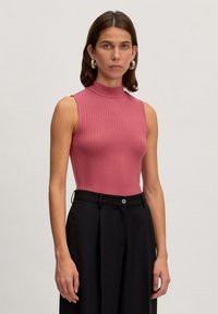 Geribbelde, mouwloze top in het roze met een hoge hals. Gemaakt in combinatie met zwarte broeken, voorzien van een klassieke knoopsluiting en een relaxte pasvorm.