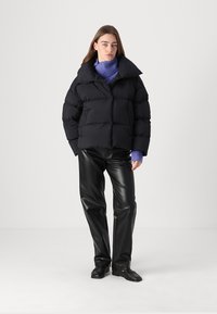 Sort puffer-jakke med høj krave, med et boksformet design og quiltet tekstur. Kombineret med glatte sorte læderbukser og en lilla sweater.