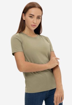 Giovane donna con lunghi capelli castani che indossa una maglietta verde oliva chiaro a maniche corte e jeans blu, posando con un braccio incrociato sull'altro.