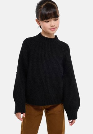 Pull en tricot noir avec un col rond, des épaules dénudées et une texture côtelée. Manches longues de style ballon. Associé à un pantalon beige.