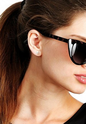 Femme aux cheveux bruns attachés en queue de cheval, portant des lunettes de soleil noires et une seule boucle d'oreille en perle blanche, regardant légèrement vers la droite.