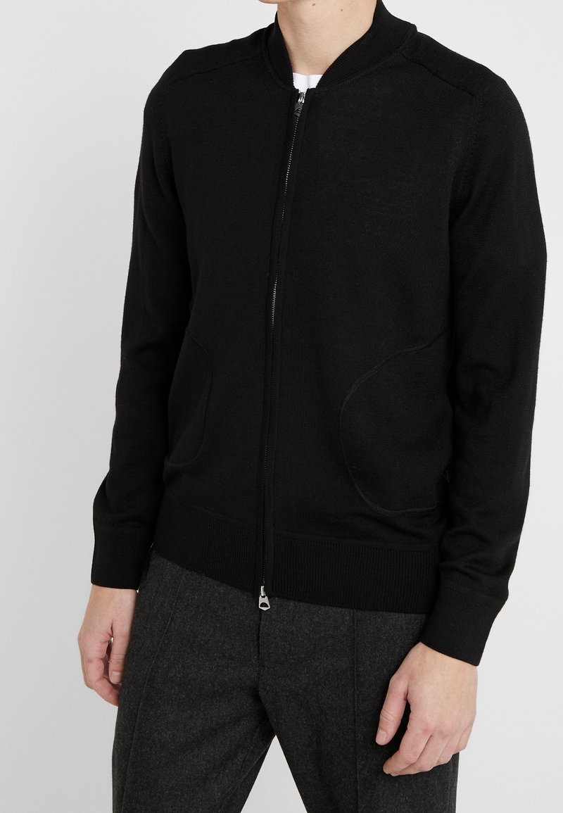 Homme portant un cardigan noir zippé avec des poches et un pantalon gris foncé sur un fond uni.