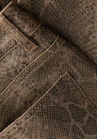 Brązowy denim w wzór wężowej skóry o fakturowanej powierzchni, z widocznym przeszyciem i detalami kieszeni, ułożony w nachodzące na siebie fałdy.