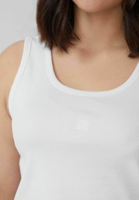 Weißes Baumwoll-Tanktop mit gerippter Struktur und rundem Ausschnitt, mit einem kleinen gestickten Logo in der Mitte.