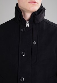 Cappotto di lana nero con colletto alto, dotato di chiusura a zip, numerosi bottoni neri e tessuto strutturato. Nessun motivo o accento visibile.