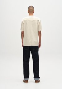 Beige Kurzarmhemd mit strukturiertem Rücken, kombiniert mit dunklen Denim-Hosen. Das Outfit zeichnet sich durch ein sauberes, minimalistisches Design aus.