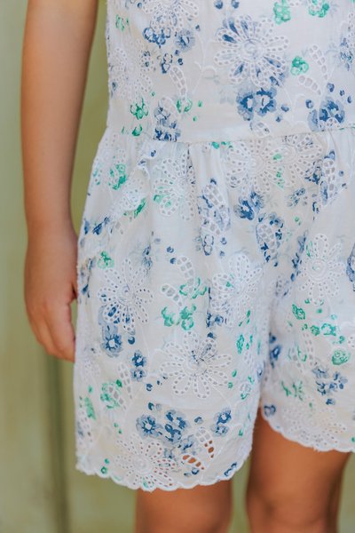 Hvit blomstrete playsuit med blå og grønne broderte mønstre, med blondekant og avslappet passform. Materialet ser lett og pustende ut.