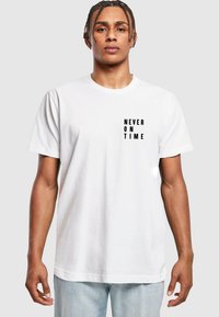 Hvid bomulds t-shirt med korte ærmer, der har sort tekst "ALDRIG TIL TIDER" på venstre side. Enkel, afslappet pasform.