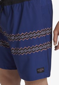 Shorts de bain bleus avec des motifs en zigzag rose et noir, avec un design de vagues. Comprend une taille élastique noire et un écusson logo.