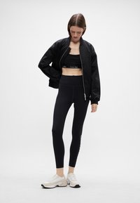 Fekete bomber dzseki egy fekete crop top és magas derekú leggings fölött, fehér logóval kiemelve. A lábbeli bézs és fehér dizájnnal rendelkezik.